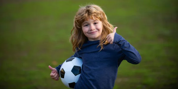 Piłkarskie dziecko gra w futbol. Dzieciak trzyma piłkę nożną, zbliżenie dzieci portret pokazać kciuki w górę znak sukcesu. — Zdjęcie stockowe
