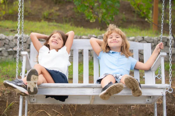 Χαριτωμένα παιδάκια που παίζουν έξω. Πορτρέτο δύο χαρούμενων παιδιών στο πάρκο. Χαριτωμένο υπέροχο αγόρι και κορίτσι αιωρείται στον κήπο ξύλινη κούνια στην πίσω αυλή. Αδερφή και αδελφός περπατούν στον κήπο. — Φωτογραφία Αρχείου
