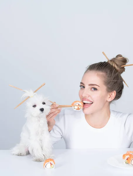 Όμορφο κορίτσι τρώει ιαπωνικά ρολά σούσι με ξυλάκια, αστείο σκυλί, παραγγείλετε στο εστιατόριο fastfood. Όμορφη γυναίκα με κουτάβι συντροφιάς κρατώντας σούσι με ξυλάκια. — Φωτογραφία Αρχείου