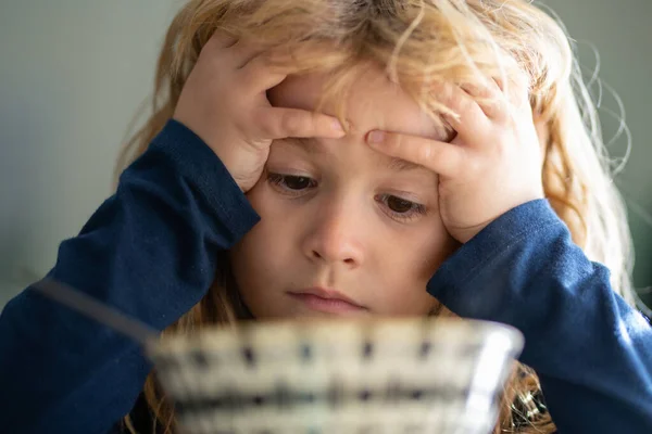 Portret van een verdrietig kind dat thuis ontbijt. Portret van een lief jongetje met blond haar, soep etend van een bord met lepel, close-up kinderen gezicht. — Stockfoto