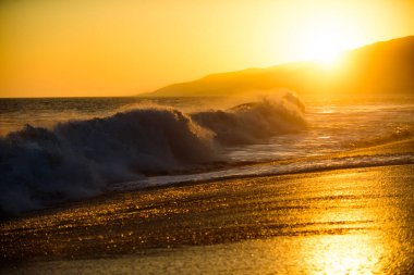 Dalgalar sıçrıyor. Altın gökyüzü ve okyanus suyu. Deniz dalgaları arka planı. Günbatımında deniz manzarası ve tropikal plaj ya da tatil ve tatil için gün doğumu. Okyanus suyu ve kumsalda kum.
