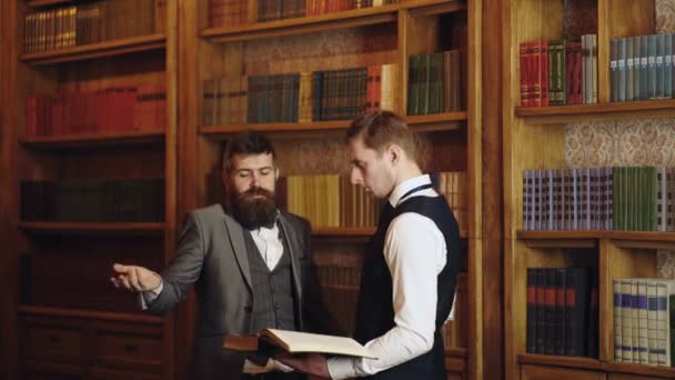 Företagsanställda chattar i biblioteket. Två män läser bok i handen på bokhyllan bakgrund. Män pratar. Begreppet lag och rättvisa. — Stockvideo