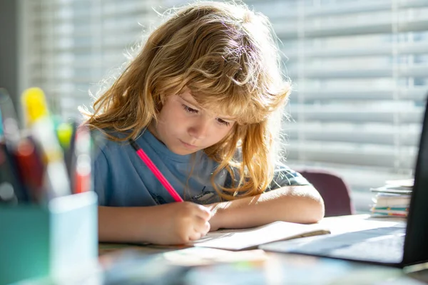 Escribir niños en la escuela. Retrato del chico de la escuela sentado en la mesa haciendo la tarea. Niño sosteniendo lápiz y escritura. Niño dibujo sobre papel blanco en la mesa. —  Fotos de Stock