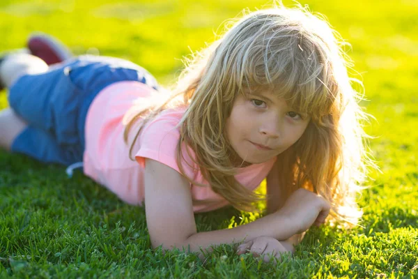 Glad liten pojke som ligger på gräset i sommarparken. Porträtt av ett leende barn som ligger på grönt gräs utomhus. Söta lilla barn på ängen i trädgården. — Stockfoto