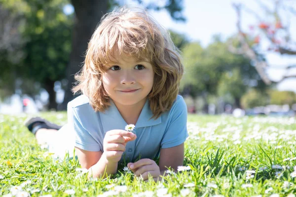 Glad liten pojke som ligger på gräset på vårdagen. Porträtt av ett leende barn som ligger på grönt gräs med tusensköna blommor i parken. — Stockfoto