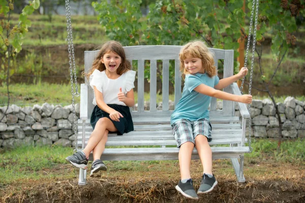 Two children swinging outdoors. Portrait of adorable brother and sister smile and laugh together while sitting on swing outdoors. Happy lifestyle kids. — Fotografia de Stock