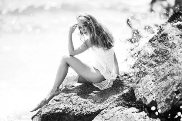 Mavi dalgalara karşı kumlu kayalıklarda oturan sakin kadının yan görüntüsü. Çekici bir genç kadın sahilde seksi bir yaz elbisesi giyiyor.. — Stok fotoğraf
