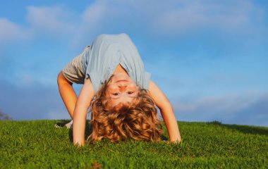 Çocuk yaz parkında yeşil çimlerin üzerinde baş aşağı duruyor. Sağlıklı aile hayatı kavramı. Eğlenceli ve tasasız bir çocukluk. Çocuklar doğada spor yapar..