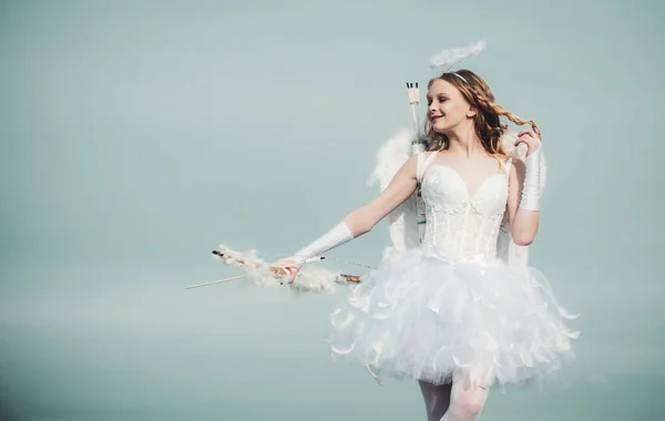 青空と白い雲に弓と矢で立つ天使の翼を持つ無邪気な女の子。ティーンエイジャーキューピッド。天使の衣装白いドレスと羽の羽を着た幼児の女の子。恋する小さな天使 — ストック写真