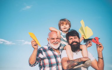 Şirin bir çocuk, babasına ve büyükbabasına sarılıyor. Mutlu gülümseyen çocuk, baba kameraya bakıyor. Mutlu üç nesil erkekler, mavi gökyüzü arka planında eğlenir ve gülümserler..