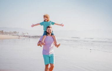 Yakışıklı adam baba, küçük oğlan çocuğu taşıyor. Mutlu baba kucağında çocuğu. Baba ve oğul yaz plajında yürüyor. Babası onu omuzlarında taşıyan küçük bir çocuk. Hafta sonu erkeği aile kavramı.