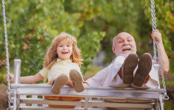 Aufgeregt schaukeln Enkelkind und Großvater im Sommergarten. Opa und Enkel amüsieren sich gemeinsam auf einer Schaukel im Sommergarten im Freien. Wochenende mit Opa. — Stockfoto
