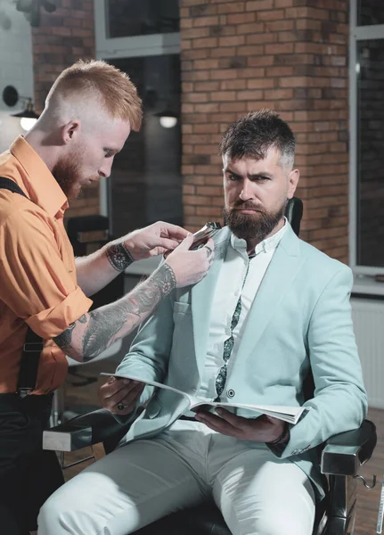 Barbeiro fazendo corte de cabelo de homem barbudo atraente na barbearia. Tão na moda e elegante. Um brutamontes na Barbearia moderna. Cabeleireiro faz penteado um homem com barba. Retrato de barba de homem elegante . — Fotografia de Stock