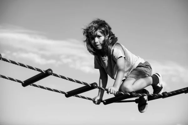 Grabben klättrar på nätet. Söt pojke klättrar upp för stegen på lekplatsen. Barn klättrar upp för stegen mot den blå himlen. — Stockfoto