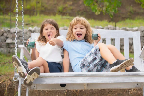 屋外で2人の興奮した子供。屋外でスイングに座っている間、愛らしい兄と妹の笑顔と笑いの肖像画。幸せなライフスタイルの子供. — ストック写真