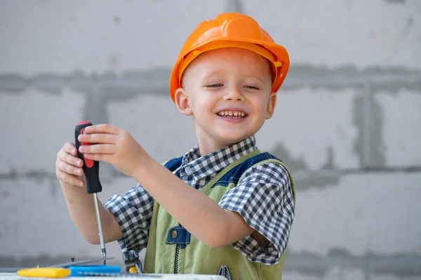 Jongetje draait bout met schroevendraaier. Kinderreparateur met reparatiewerktuig. Kind in helm en boilersuit op bouwplaats. Kleine arbeidersingenieur. — Stockfoto
