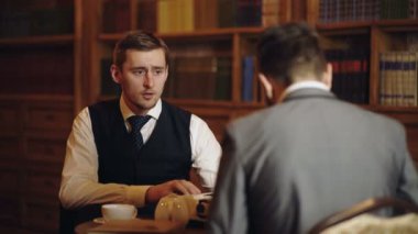 İş adamları kütüphanede sohbet ediyor. Profesör klasik kütüphane dolabında kitap okuyor. Takım elbiseli adamlar dolapta bir şeyler konuşuyor ya da tartışıyorlar. Erkekler konuşuyor ve tartışıyor.