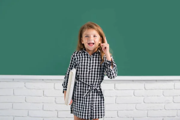 Portrait of school girl nerd student hold book with surprising expression pointing with finger against blackboard. Back to school. — Stok fotoğraf