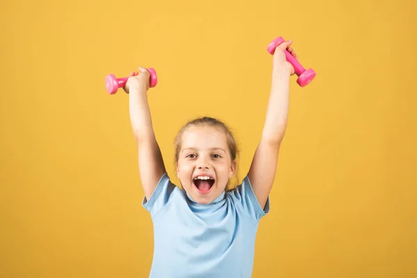 Child girl is doing exercises with dumbbells on yellow studio background. Sport portrait kids. — Stockfoto