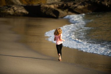 Deniz sahilinde koşan mutlu çocuk. Komik çocuk sörf kenarına doğru koşuyor. Aktif çocuk yaşam tarzı. Eğitimdeki küçük bir atlet. Koşucu spor yapıyor, çocuklar için koşuyor..