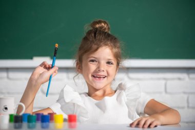 Child girl draws in classroom sitting at a table, having fun at school blackboard background. Painting school lesson, drawing art.