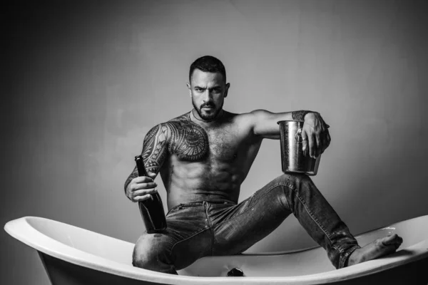 Hombre guapo barbudo sin camisa en jeans con cuerpo sexy en el baño. Hombre macho sexual en el baño. Fuerte musculoso hombre tatuado sosteniendo botella de champán y posando en el baño . — Foto de Stock