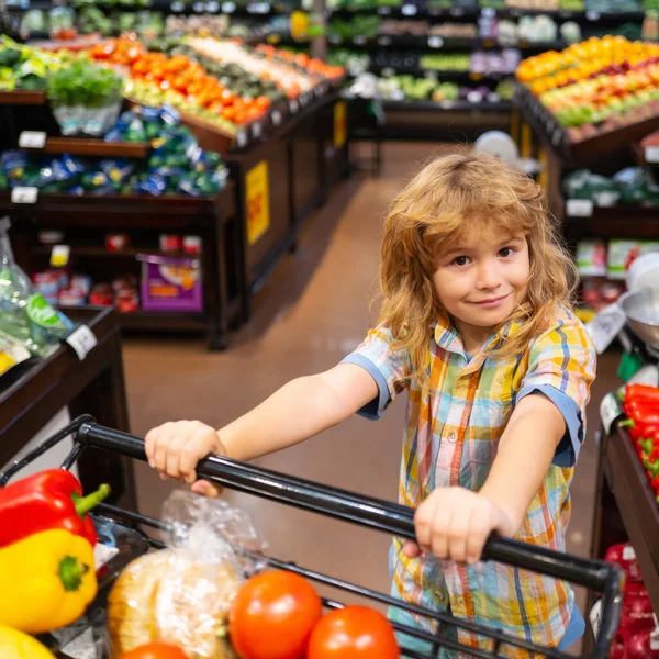 Liten söt pojke med kundvagn full av färska ekologiska grönsaker och frukter i mataffär eller snabbköp. Pojke ha kul när du väljer mat i snabbköpet. — Stockfoto