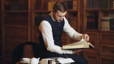 Portrait of authentic man reading book in the hand on bookshelf background. Handsome well-dressed man stands by bookshelves in a room with classic interior. Old fashion.
