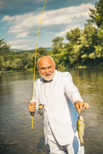 一个快乐的老人钓鱼的画像。爷爷有鱼吃成熟的渔夫,身穿白色西服,打着钓竿,在河里纺丝.捕鳟鱼. — 图库照片