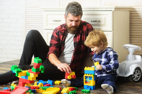 Father takes part in game with son playing on floor holding toy cars. Father and son create toys from bricks. Dad and kid build of plastic blocks. Generations family.