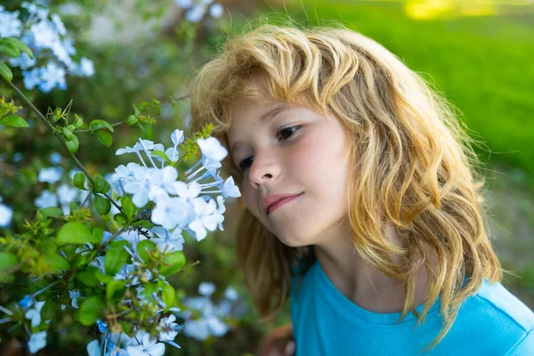 Tavaszi gyerek arc. Közelkép egy kis szőke gyerekről tavaszi virággal. Vicces kölykök a kertben. A gyerekek összezárják arcukat a virágos háttérrel. Gyermek lány játszani tavaszi virágok. — Stock Fotó