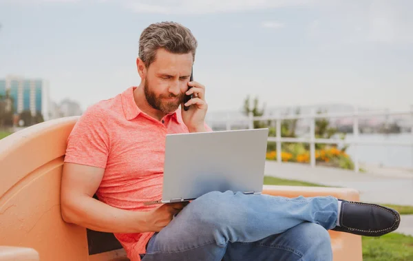 Snygg ung affärsman som arbetar med bärbar dator utomhus pratar på mobiltelefon. — Stockfoto