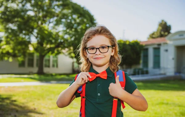Alegre niño de escuela en uniforme escolar con mochila de pie cerca de la escuela. De vuelta a la escuela. Concepto de educación preescolar infantil. —  Fotos de Stock
