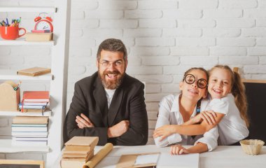Baba, küçük ilkokul öğrencilerine, kızların okul ödevlerini hazırlamalarına, masada yan yana oturmalarına yardım ediyor. Mutlu küçük çocuklar babalarıyla evde eğitim görüyor..