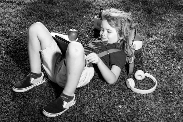 Kind sitzt mit Tablet auf Gras im Park. Schule im Freien. Lächelnder Junge hält Tablet-Computer in der Hand. Schule, Bildung, Lernen, Technologie Freizeit Outdoor-Konzept. — Stockfoto