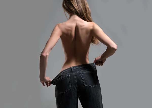 Slim girl have points at slim waist in big trousers, successful weight loss, isolated on gray background. Successful weight loss, woman with too large jeans after a diet. Skinny waist, back view.