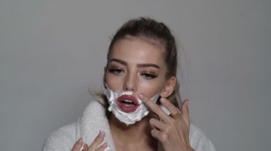 Çekici genç bir kadın yüzünü tıraş ediyor. Neşeli komik kadının yüzünde krem var. Kendini jiletle tıraş ediyor. Trans cinsiyet kavramı.