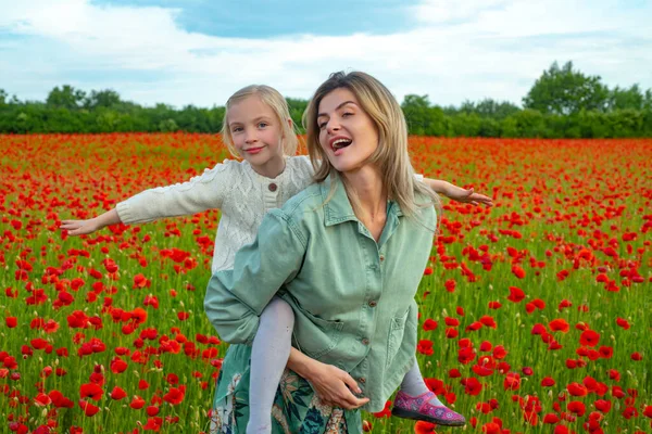 Mother and daughter walking through a flowering poppy field. Child and mom piggyback ride on summer poppies meadow with poppys flowers. Happy motherhood. Spring design. —  Fotos de Stock