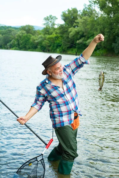 Pescatore che pesca su un rever. Buon vecchio pescatore catturato un pesce trota. Ritratto di allegro sorridente uomo anziano pesca. Nonno con pescato. — Foto Stock