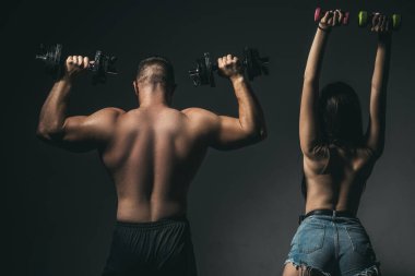 Çift halterlerle birlikte egzersiz yapıyor. Çıplak vücutlu kaslı bir adam, arka planda koyu renkli halterleri olan fitness kadını. Seksi, formda bir vücut. Dambılı çift eğitimi.