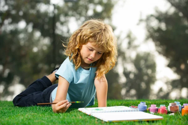 Kid draws in park laying in grass having fun on nature background. Children artist paints creativity vacation, kids crafts. Stock Photo