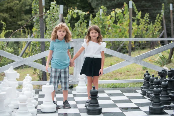 Testvérek nagy sakktáblán játszanak. Kisfiúk és kislányok élvezik a tavaszt. Gyerekek sétálnak az udvaron. Legjobb barátok. Két gyerek sakkozik.. — Stock Fotó
