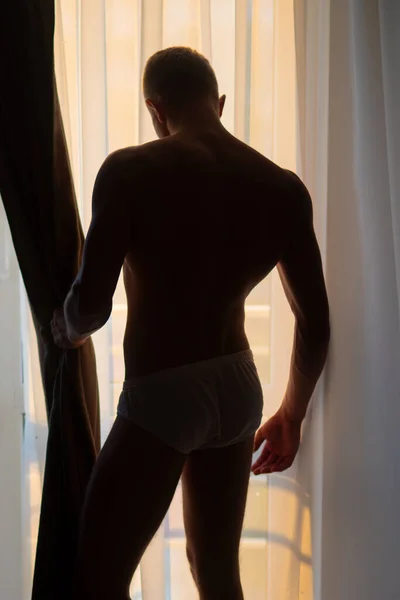 Pensativo y guapo joven sin camisa mirando por una ventana. Sexy silueta gay en ventana sombra. —  Fotos de Stock