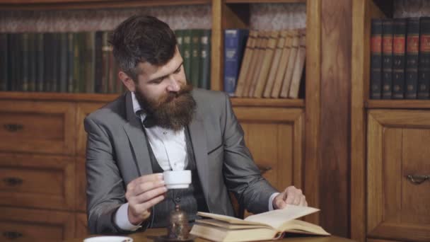 Зріла людина в бібліотеці і насолоджується читанням. Бородатий чоловік у костюмі виглядає задоволеним. Розслабтеся, насолоджуйтеся відпочинком, концепцією хобі. Література та успіх. Пийте каву або чай. Професор . — стокове відео