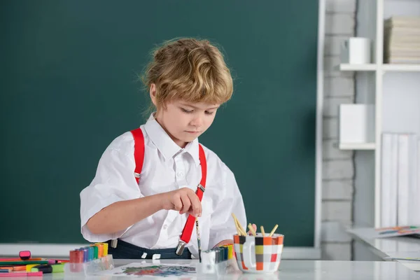 A kisfiú rajzol az osztályteremben ül az asztalnál, és szórakozik az iskola táblás hátterén. Gyerekek korai művészet és kézművesség oktatás. — Stock Fotó