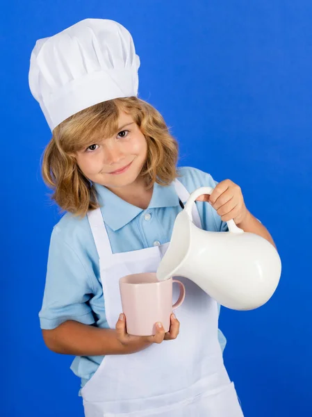 Çocuklar için süt. Çocuk aşçı, izole edilmiş mavi stüdyoda yemek hazırlıyor. Çocuklar yemek yapıyor. Önlüklü ve şapkalı genç çocuk mutfakta sağlıklı bir sebze yemeği hazırlıyor.. — Stok fotoğraf