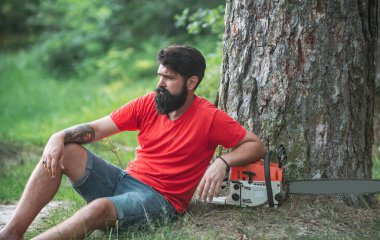 Ormanın yakınında baltası olan yakışıklı bir genç. Elinde elektrikli testereyle oduncu. Elektrikli testere. Ormanların tahrip edilmesi doğal ekosistemlerin bozulması ve istikrarsızlaşmasının başlıca nedenlerinden biridir..