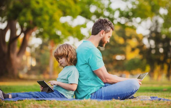 Enseignement à distance extérieur. Père utilisant ordinateur portable se détendre avec le fils écolier tenant ordinateur portable s'amuser ensemble. Famille avec gadgets à l'extérieur sur la nature. — Photo