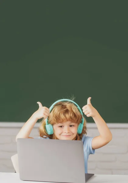 Rapaz bonito com polegares para cima usando fones de ouvido, estudar com laptop em sala de aula, ouvir curso de aula de áudio. Primeiro dia na escola. — Fotografia de Stock