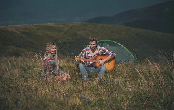 Καταπληκτικό νεαρό ζευγάρι αγάπης στην κατασκήνωση έξω σε δωρεάν εναλλακτικές διακοπές κάμπινγκ πάνω από τα βουνά. Ευτυχισμένοι ταξιδιώτες να ξεκουραστούν και να παίξουν κιθάρα. Ενεργός τρόπος ζωής και καταπληκτική έννοια βουνά. — Φωτογραφία Αρχείου